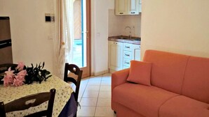 A dining area with a table set for two, a sofa, and a kitchen in the background. - Appartamenti Granseola - Granseola -  Bilocale (Campo nell'Elba)