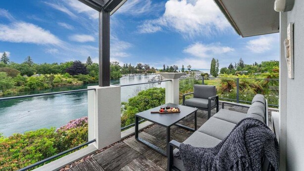 A balcony with a view of a river and greenery, a table with a plate of food, and a sofa with a blanket. - The Pier Taupo (Taupo)