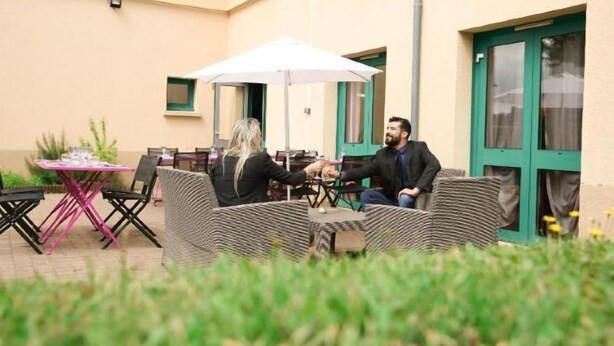 An outdoor seating area with a table and chairs, a white umbrella, and a building with green doors. - Hôtel Inn Design Resto Novo Châteaubriant (Chateaubriant)