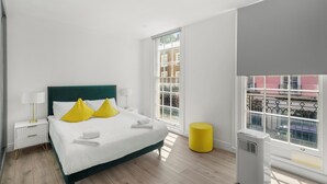 A modern hotel room with a large bed, white bedding, a green headboard, and a yellow ottoman. - Flat 1 Beauchamp Place (London)