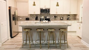 A kitchen with white cabinets, a granite countertop, stainless steel appliances, and gold bar stools. - Themed 5Bd Home, Movie and Game Room (4726) (Kissimmee)