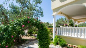 Un giardino con una pergola ricoperta di fiori rosa, un vialetto lastricato e un balcone bianco con piante in vaso.