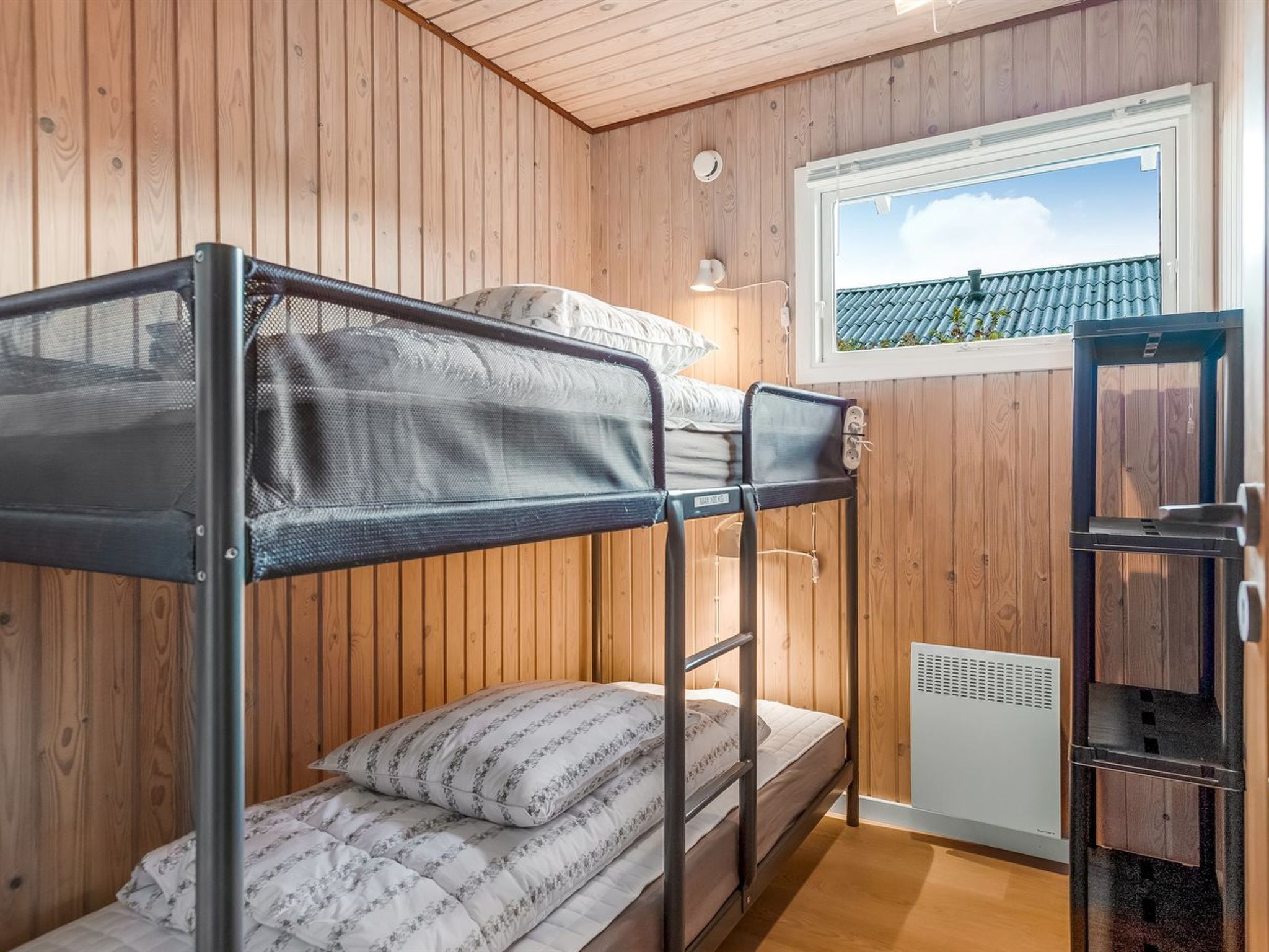 A bunk bed room with wooden walls, a window with a view of a tiled roof, and a white heater.