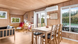 Een gezellige kamer met een houten plafond, een eettafel met stoelen, een rode fauteuil, een bank en een raam met uitzicht op groen.