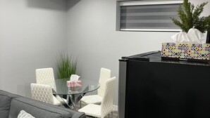 A modern living room with a grey sofa, a black refrigerator, and a dining area with white chairs and a glass table.