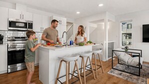 A modern kitchen with white cabinetry, a central island with stools, stainless steel appliances, and a staircase leading to an upper level. - Luxury Family Retreat w/ Games, King Bed & Garage (Edmonton)