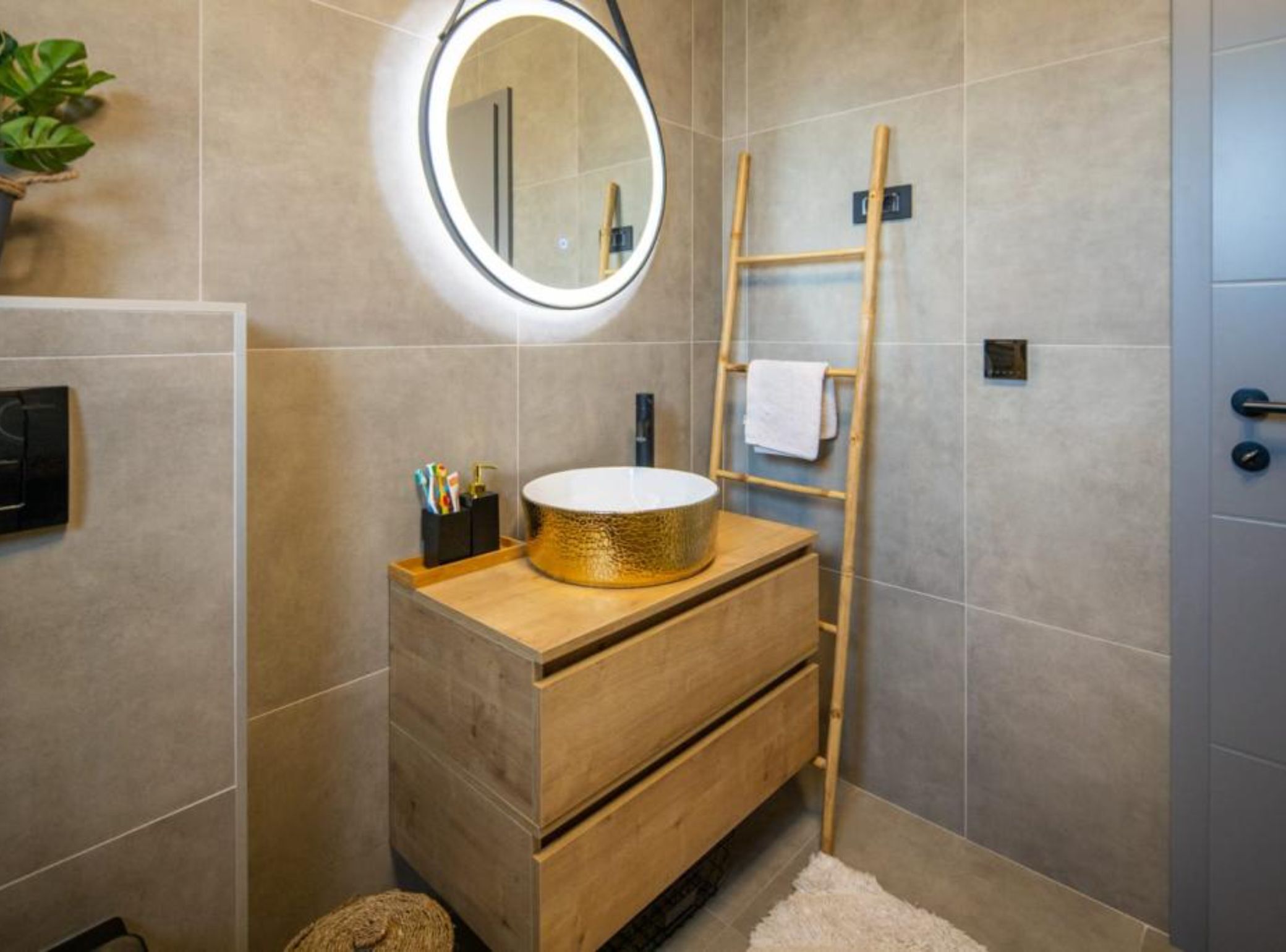 Een badkamer met een houten wastafelmeubel, een ronde spiegel, een handdoekrek en een kleine plant.