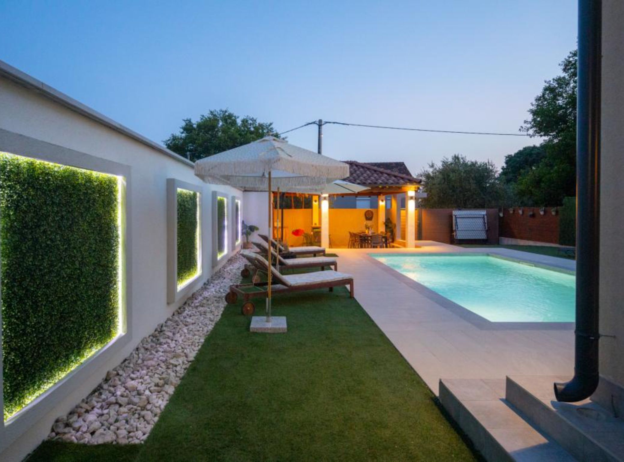 Ein moderner Poolbereich im Freien mit Liegestuhl und Sonnenschirm, umgeben von einer gepflegten Rasenfläche und einer Wand mit belebten Pflanzen.