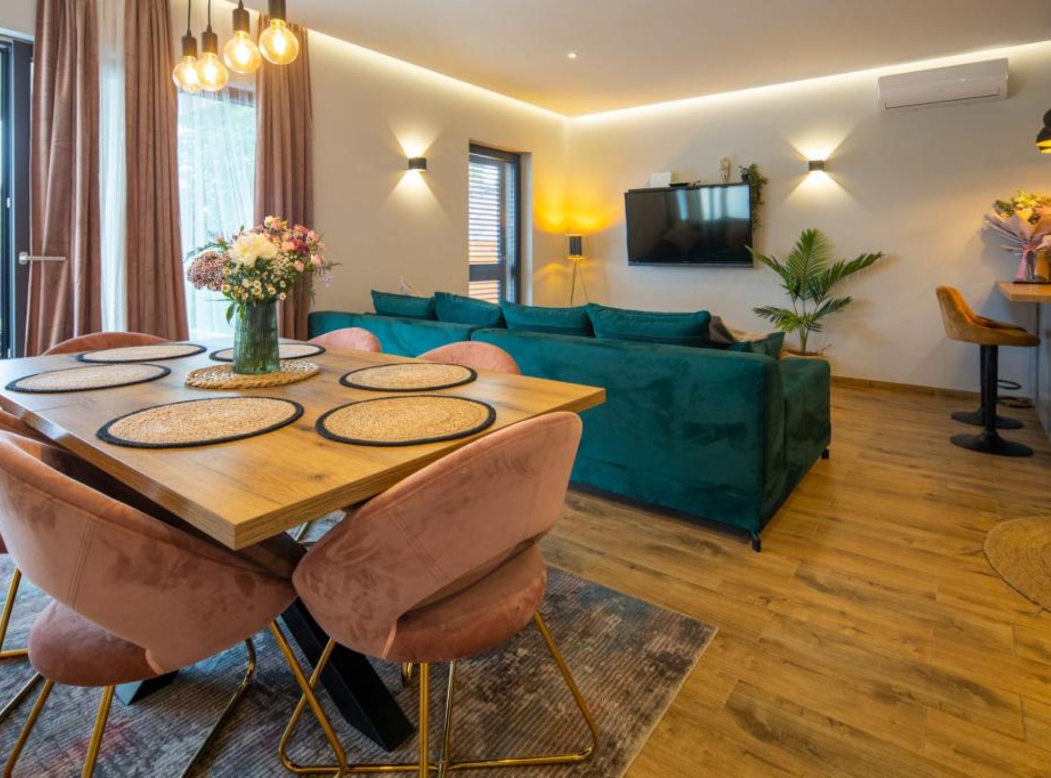 Una sala moderna con una mesa de comedor de madera, sillas de terciopelo rosa, un sofá color turquesa y un televisor de pantalla plana montado en la pared.