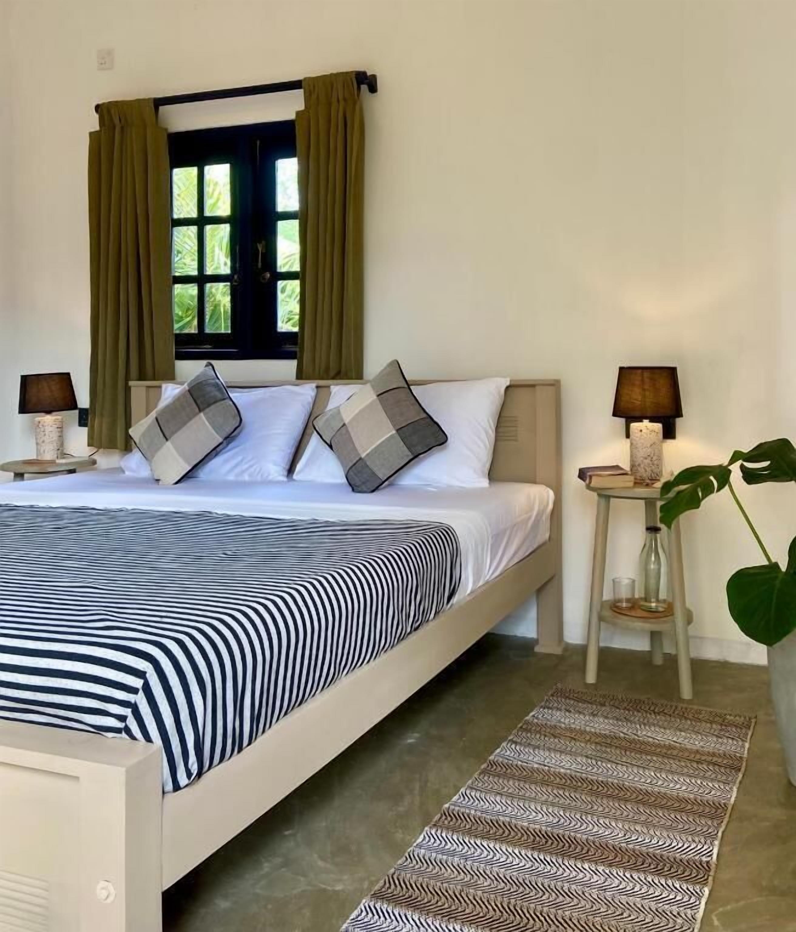 A neatly made bed with striped bedding, two bedside lamps, a window with curtains, and a patterned rug.