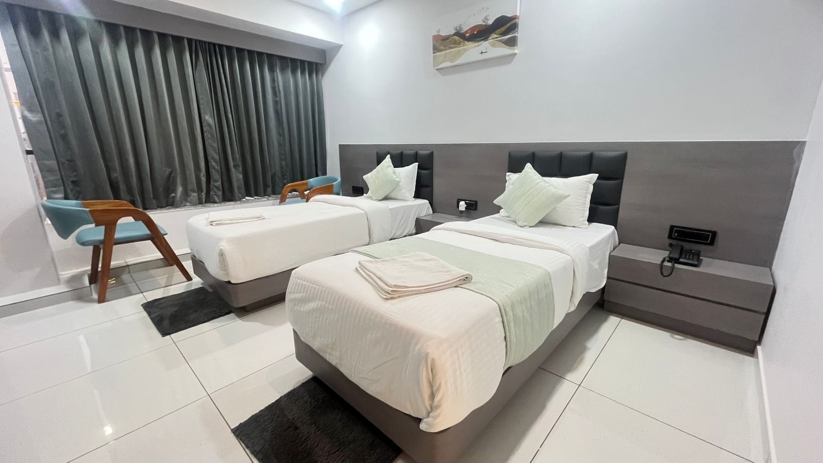 A modern hotel room with two beds, a nightstand, a chair, and a painting on the wall.