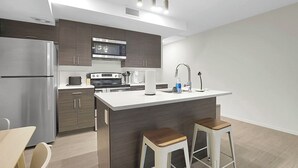 Een moderne keuken met een roestvrijstalen koelkast, magnetron en kookplaat, een wit aanrecht en twee houten krukken.