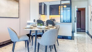 Un espace repas moderne avec une table ronde pour quatre personnes, entourée de chaises à revêtement gris. On aperçoit à l’arrière-plan une cuisine avec des meubles sombres et une fenêtre.