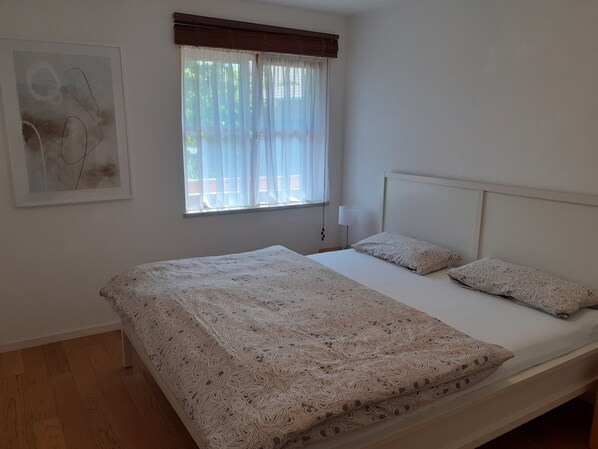 Ein Schlafzimmer mit einem Einzelbett, einem Fenster mit leichtem Vorhang, einer Nachttischlampe und einem gerahmten Bild an der Wand.