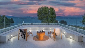 Eine Terrasse mit einem runden Holztisch, zwei Stühlen und einem weißen, flauschigen Sessel. Im Hintergrund steht ein großer Baum, und die Terrasse bietet Blick auf ein Gewässer.