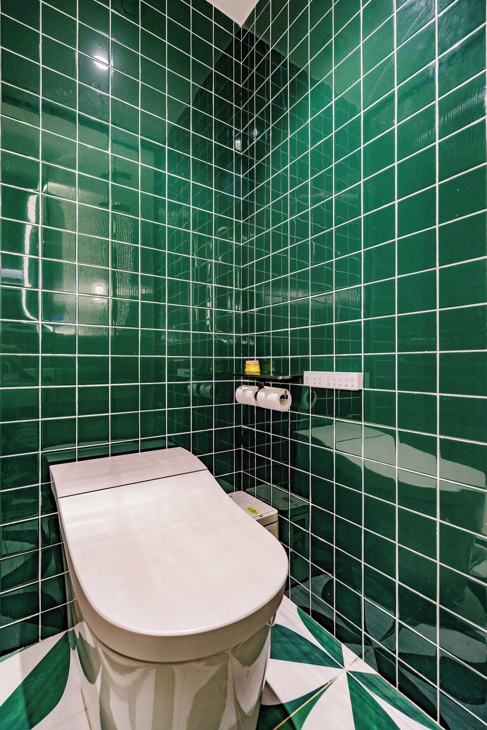 A bathroom with green tiled walls, a white toilet, and a paper towel dispenser.