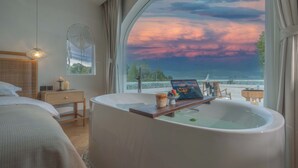 Un dormitorio con una bañera grande, una mesita de noche con velas y vistas a un atardecer sobre el agua.
