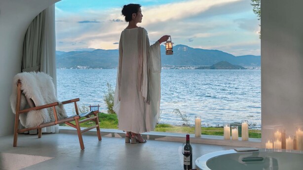 Una persona vestida de blanco sostiene un farol, de pie junto a una ventana con vistas a una masa de agua y montañas al fondo.
