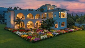 Un edificio de varios pisos con ventanas arqueadas y un balcón, rodeado de un jardín cuidado con diversas flores.