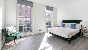 A bedroom with a large bed, a green chair, and a view of a pink building through a window. - Flat 2 Beauchamp Place (London)