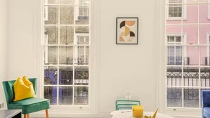 A room with large windows, a green chair with yellow cushions, a framed abstract artwork, and a table with a yellow object. - Flat 2 Beauchamp Place (London)