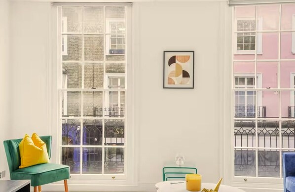 A room with large windows, a green chair with yellow cushions, a framed abstract artwork, and a table with a yellow object. - Flat 2 Beauchamp Place (London)