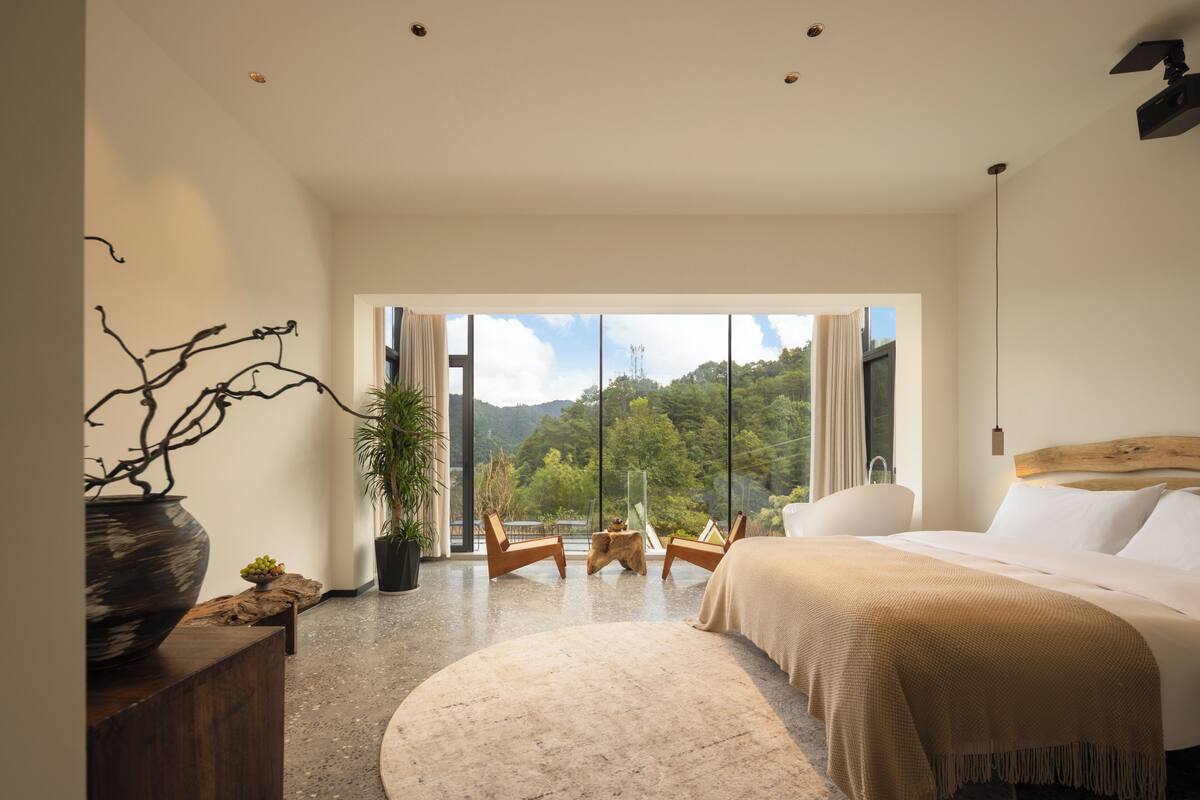 A modern bedroom with a large bed, a view of the outdoors, and a wooden side table with a vase.