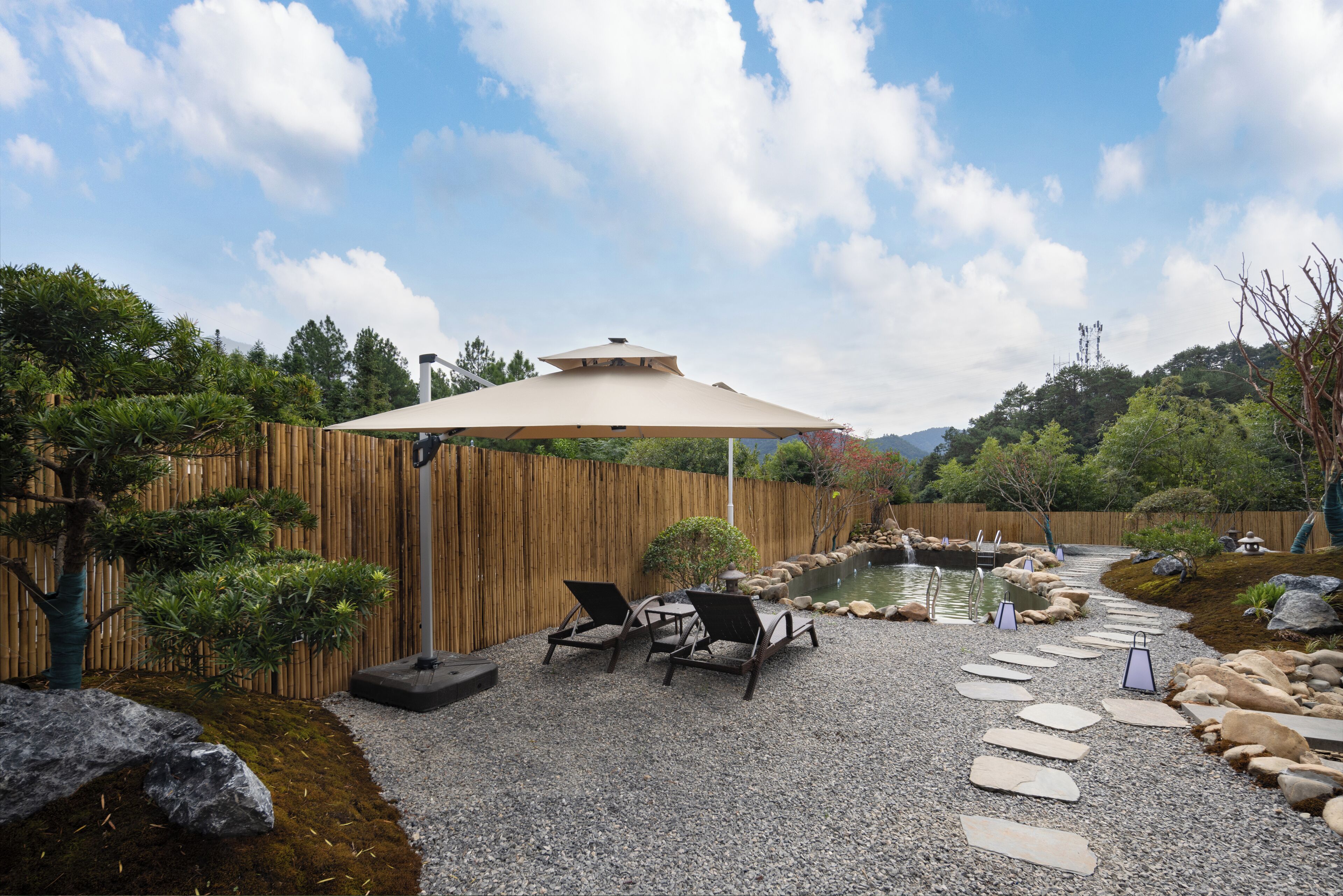 A serene outdoor area with a wooden fence, a koi pond, and a stone pathway leading to two lounge chairs under a large umbrella.