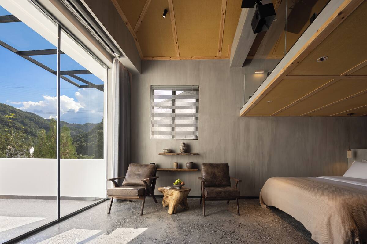 A modern bedroom with a large window, two leather chairs, a wooden coffee table, and a bed.