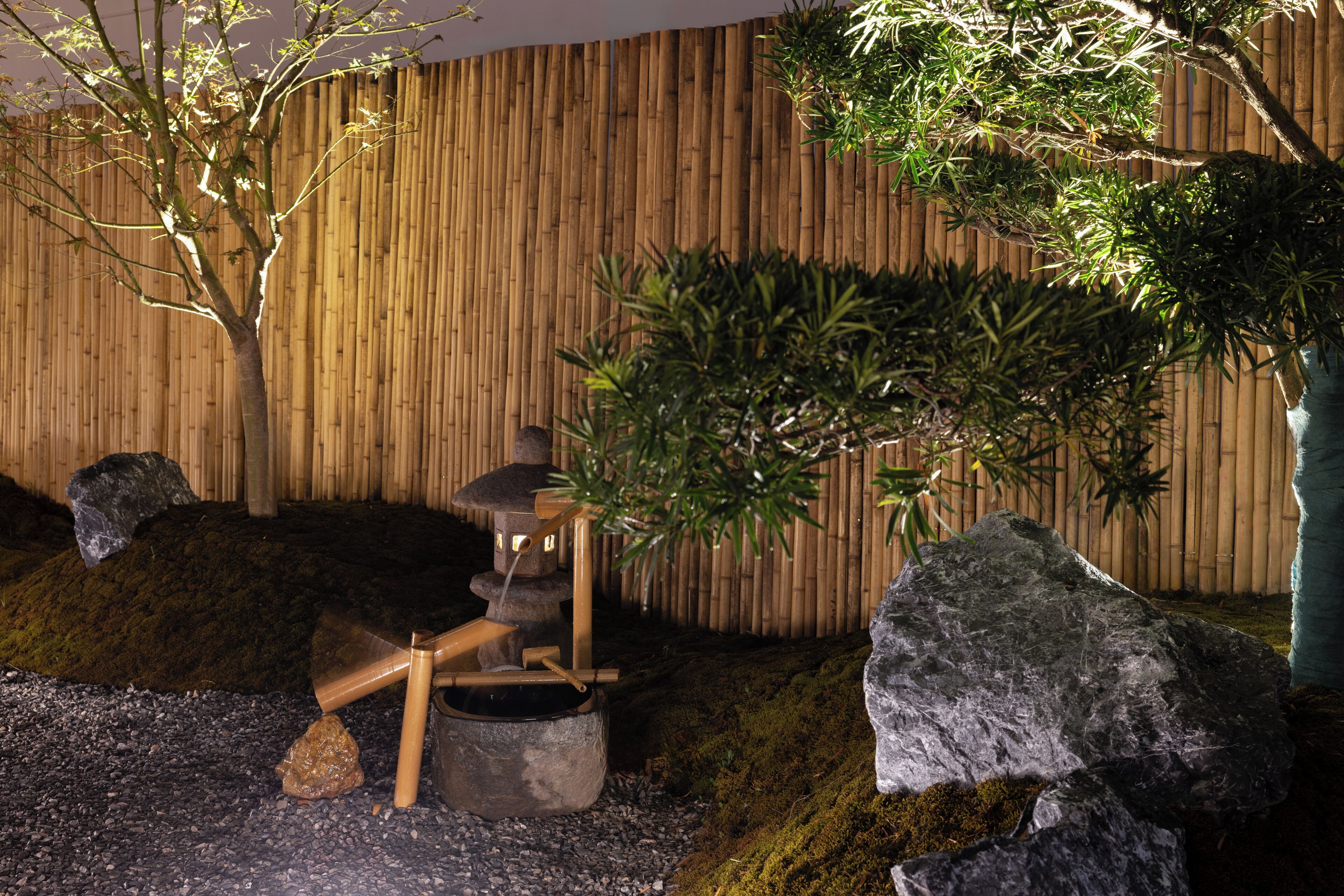 Un jardín de rocas con cerca de bambú, un elemento acuático y un árbol.