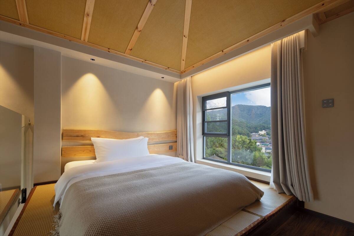 A bedroom with a wooden headboard, a large bed with white bedding, a view of a mountainous landscape through a window, and a wooden ceiling.