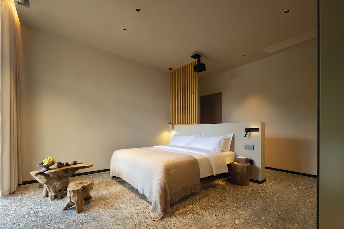 A modern hotel room with a large bed, a wooden side table, and a small round table with a bowl of fruit.