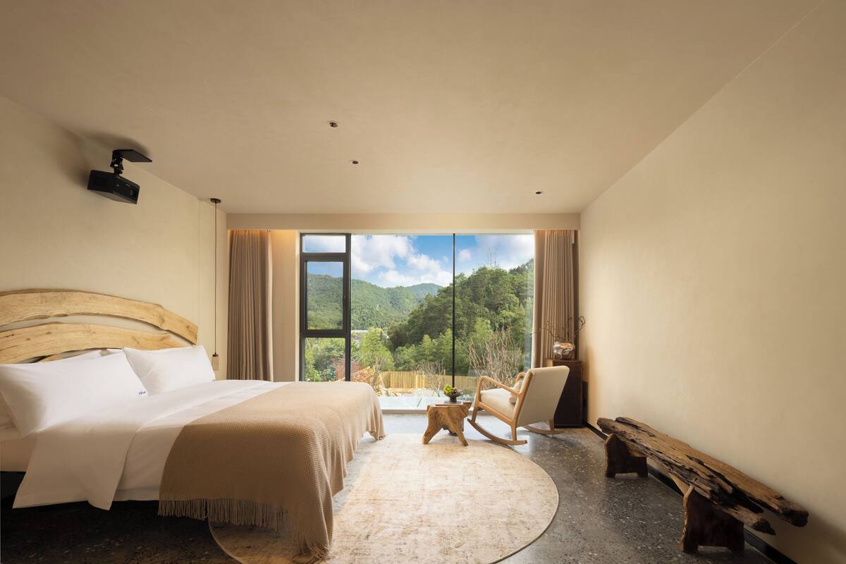A bedroom with a large bed, a wooden rocking chair, a small table, and a bench.