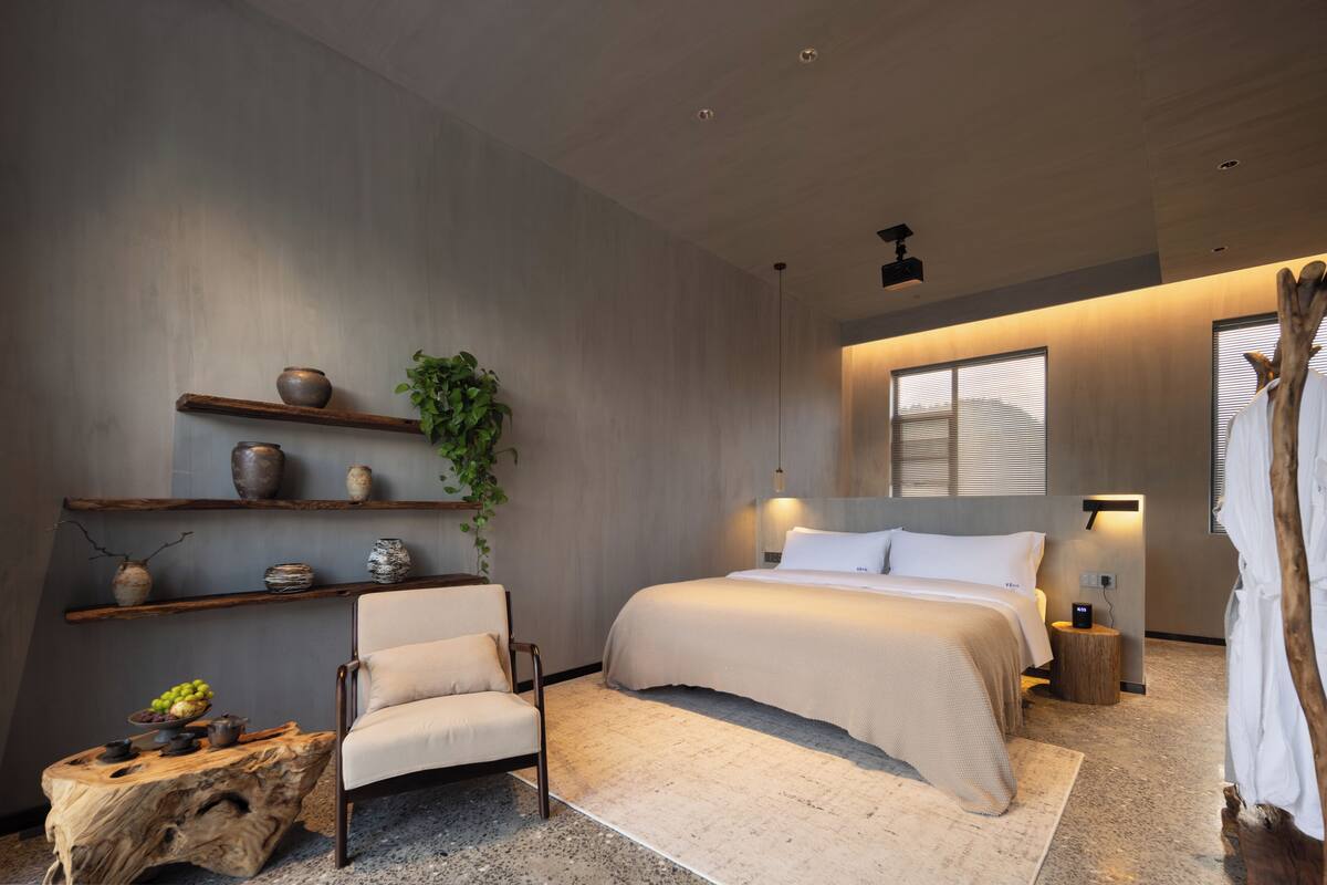 A modern bedroom with a large bed, a chair, a wooden side table, and shelves with decorative items.