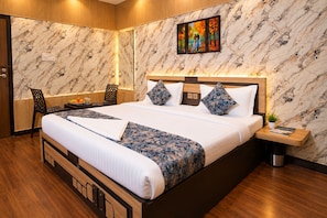 A hotel room with a large bed, wooden headboard, marble-patterned walls, a painting above the bed, a small side table, and a chair. - Hotel Kasol Heritage (Lucknow)