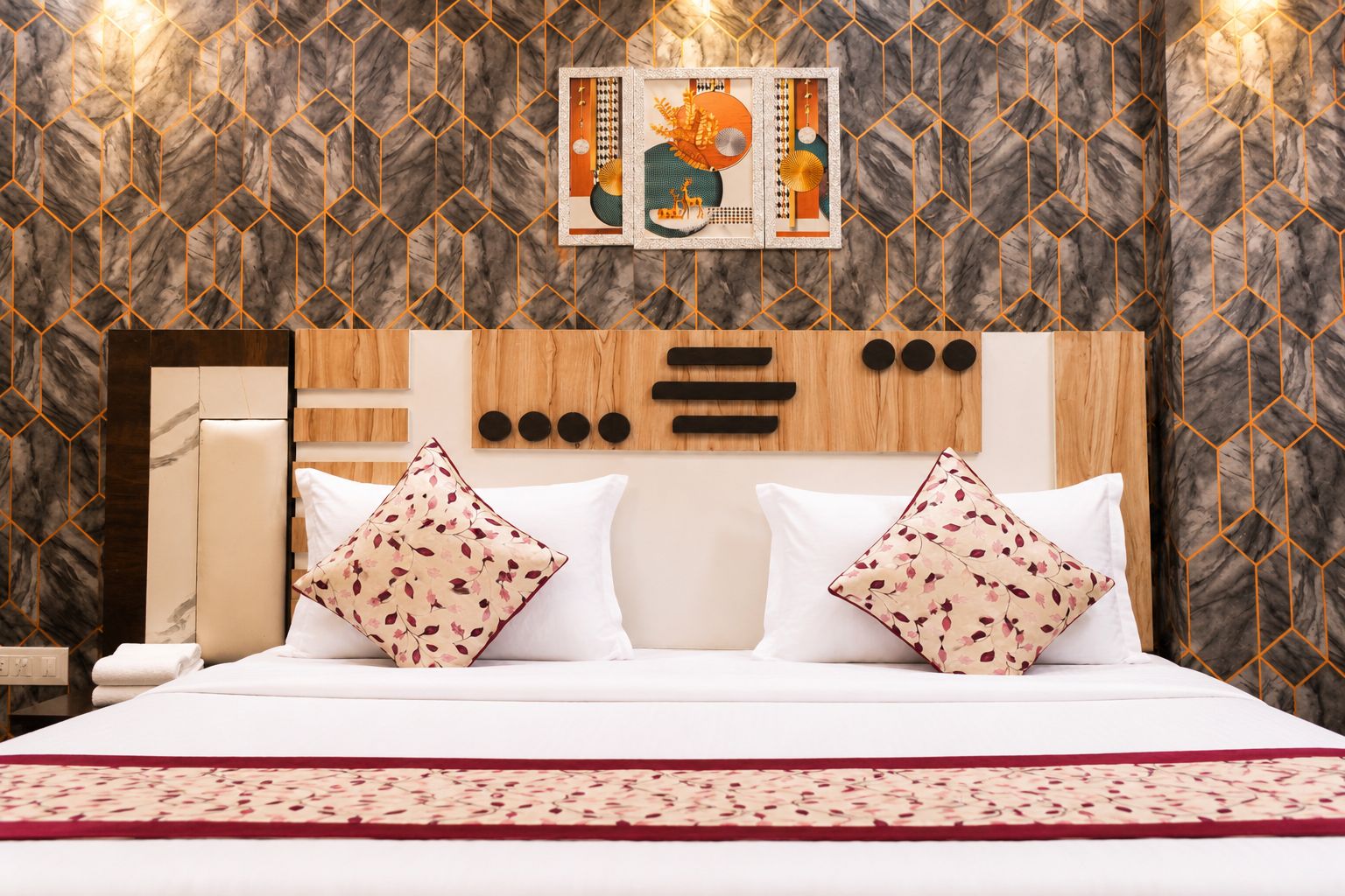 Ein Hotelzimmer mit Wand in Marmoroptik, einem hölzernen Kopfteil, zwei weißen Kissen mit Blumenmuster und einer roten und weißen Tagesdecke.