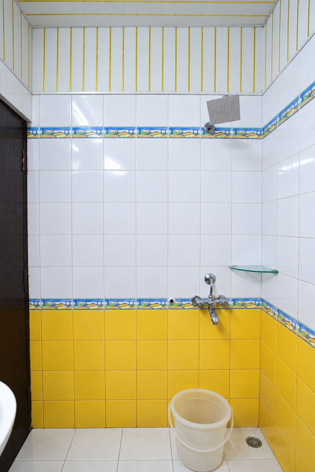 Une salle de bain avec des carreaux jaunes et blancs, une douche, une étagère en verre et un seau.