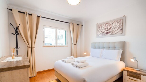 A neatly arranged bedroom with a bed, bedside tables, a window with curtains, a coat rack, and a wall-mounted artwork. - GuestReady - Machico Apartment with Balcony (Machico)