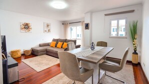 A modern living room with a sofa, dining table, and chairs. There is a TV stand and a window with curtains. - GuestReady - Machico Apartment with Balcony (Machico)