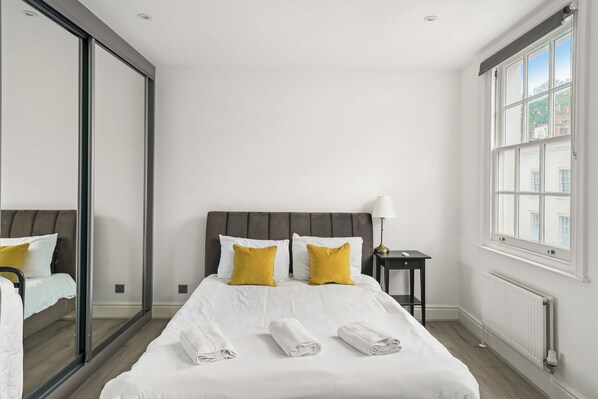 A hotel room with a large bed, two pillows, a nightstand, a lamp, and a window. - Flat 3 Beauchamp Place (London)