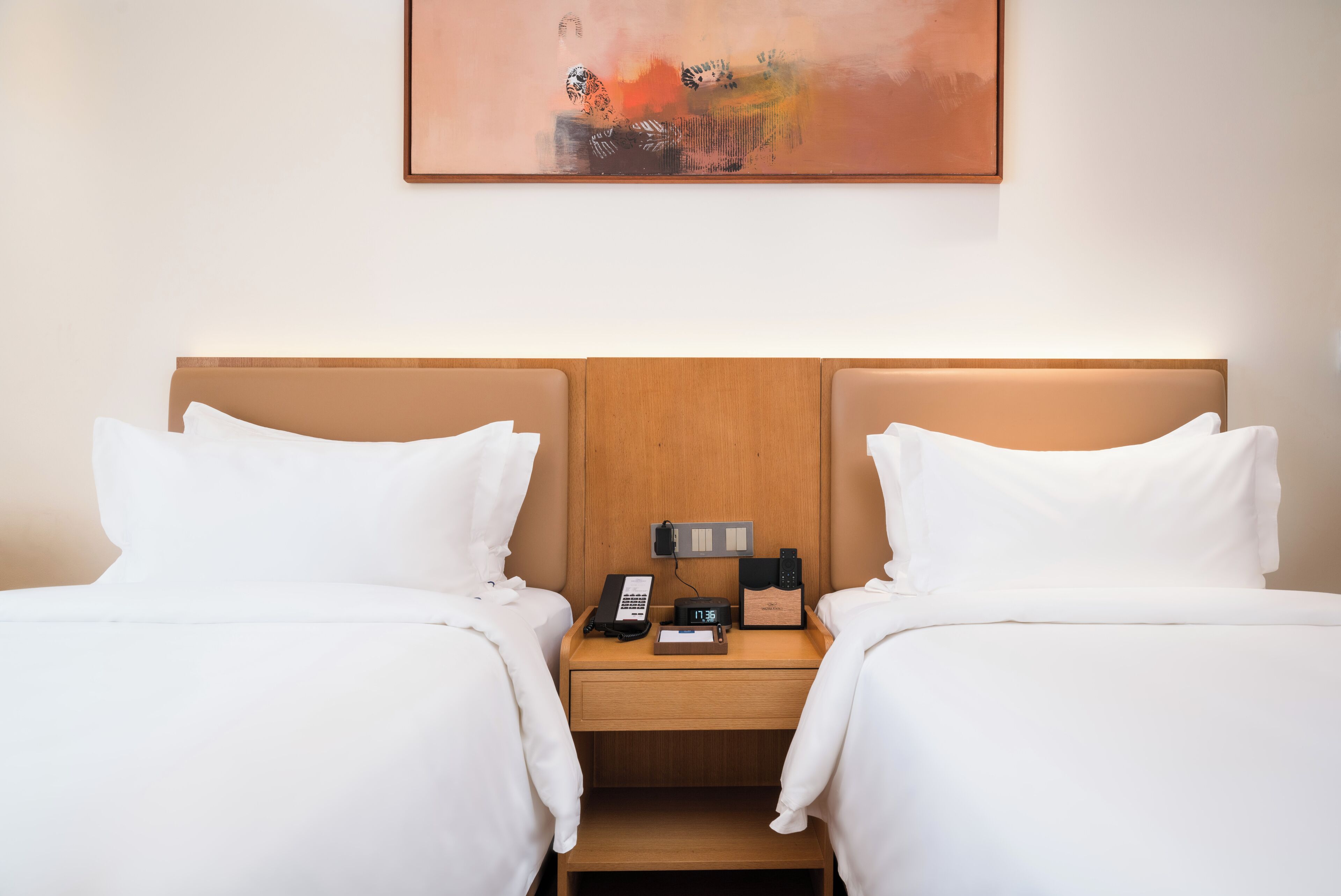 A hotel room with two beds, a nightstand with a phone and a clock, and a painting on the wall.