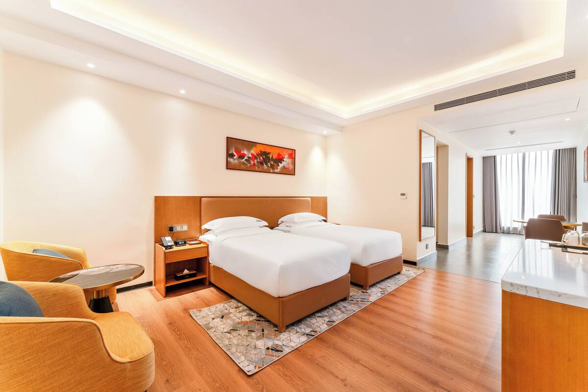 A hotel room with a bed, bedside table, lamp, chair, and a view of the dining area.