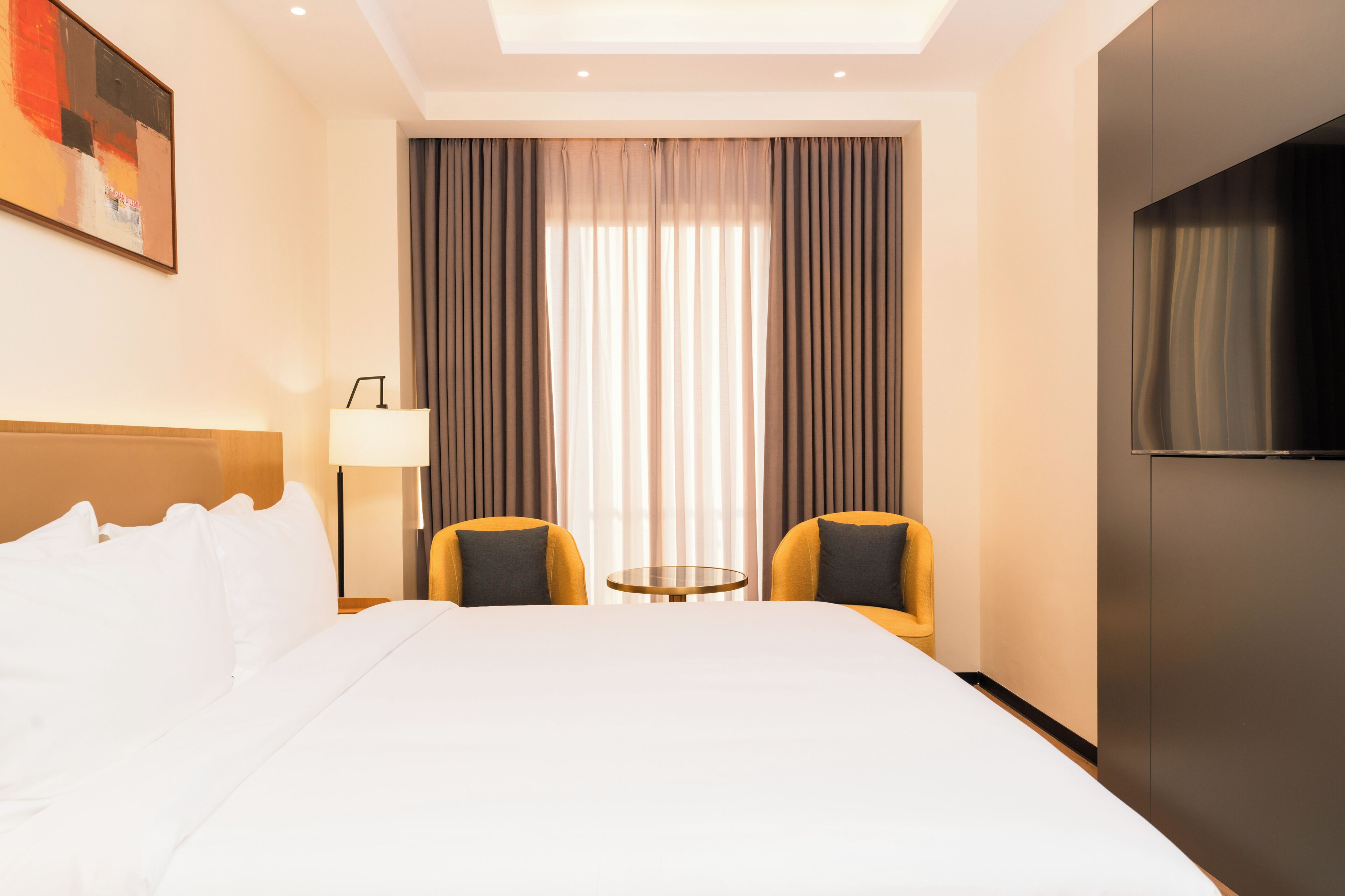 A hotel room with a bed, two chairs, a small table, a TV, and a window with curtains.