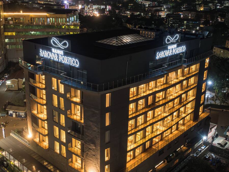 A multi-story building with illuminated signage for 'BASIL SAROVAR PORTICO' and a rooftop terrace.