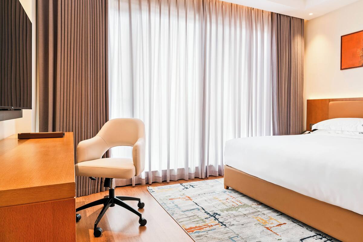 A hotel room with a bed, a desk with a chair, a TV, and a large window with curtains.