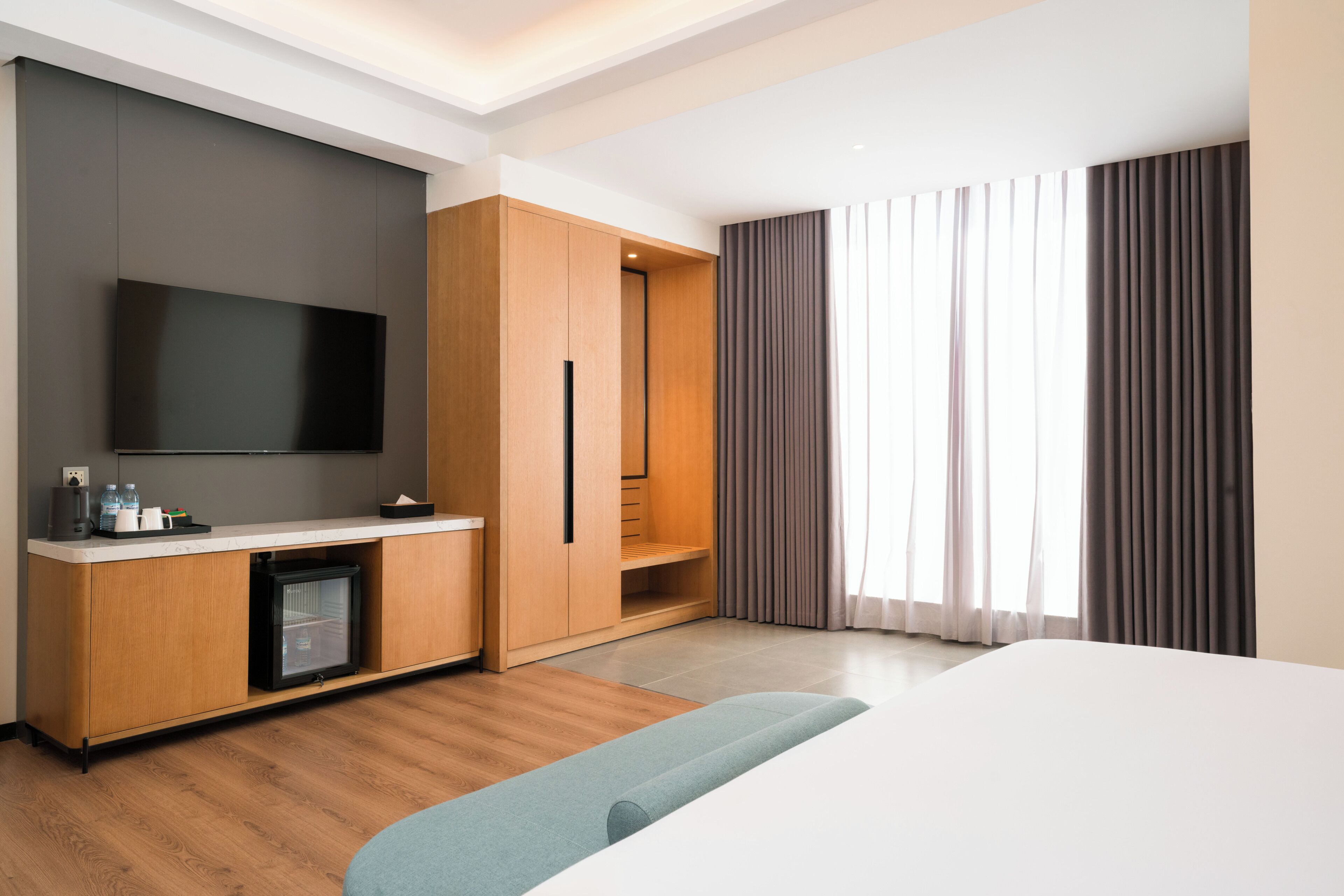 A modern hotel room with a flat-screen TV, a minibar, a wooden wardrobe, a seating area, and a large window with curtains.