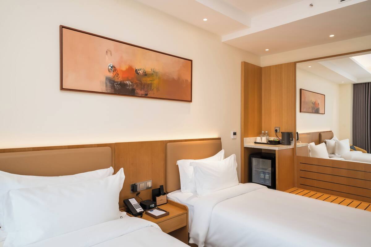 A hotel room with two beds, a nightstand with a phone and a coffee maker, a small refrigerator, and a painting on the wall.