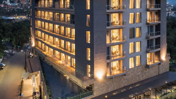 Un hotel de varios pisos con ventanas y balcones iluminados, un letrero que dice 'SAROVAR PORTICO' y vistas de la ciudad circundante.