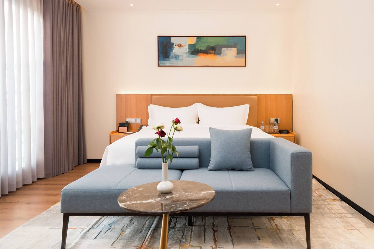 A modern hotel room with a bed, a sofa, a small table, and a painting on the wall.