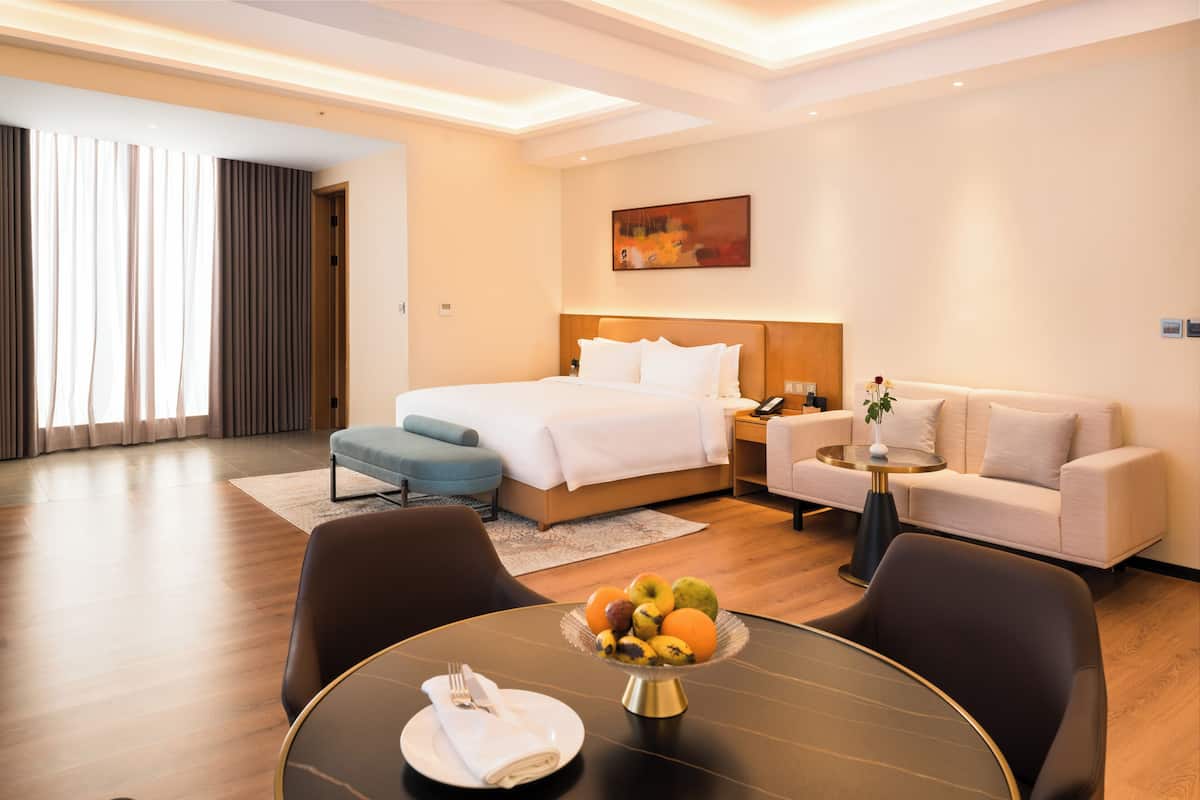 A hotel room with a bed, a sofa, a dining table with fruit, and a painting on the wall.